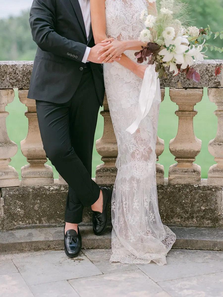 Bride in a lace gown and groom in a black suit by a château's stone balustrade
